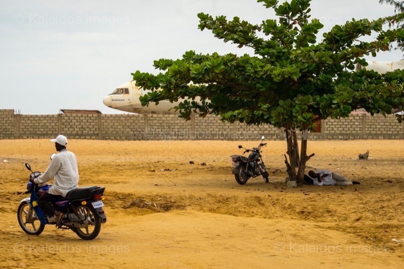 Barakah, L1011 TriStar, Lockheed L1011, abandoned TriStar, aircraft wreck, grounded aircraft, white fuselage, derelict cockpit, empty cabin, Cotonou beach, Cadjéhoun beach, Cotonou Benin, West Africa, Benin airport, Atlantic coastline, coastal Benin, corroded wreck, marine rust, deterioration, salt corrosion, decay, abandonment, dereliction, dilapidation, environmental erosion, aviation heritage, industrial archaeology, failed tourism project, open-air museum, urban art, colonial relic, Benin history, alternative tourism, architectural curiosity, aviation history, hajj pilgrimage, Air Rum airline, defunct carrier, humanitarian transport, international migration, historical traces, industrial heritage, stranded aircraft, tourist attraction, urban exploration, ruin photography, beach adventure, Beninese leisure, amusement park, immersive experience, urban safari, overcast sky, sunset beach, tropical storm, coastal erosion, sea salt, Atlantic Ocean, white sand, coastal vegetation, gleaming white, nature-machine contrast, aerospace geometry, outstretched wings, empty windows, cabin stairway, cockpit instruments, time displacement, obsolescence, suspended time, liminality, aborted transformation, urban memory, unfinished dream, nature-industry symbiosis, tourism ghost, frozen history