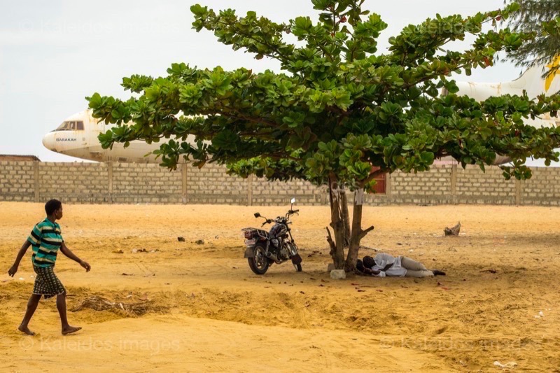 Barakah, L1011 TriStar, Lockheed L1011, abandoned TriStar, aircraft wreck, grounded aircraft, white fuselage, derelict cockpit, empty cabin, Cotonou beach, Cadjéhoun beach, Cotonou Benin, West Africa, Benin airport, Atlantic coastline, coastal Benin, corroded wreck, marine rust, deterioration, salt corrosion, decay, abandonment, dereliction, dilapidation, environmental erosion, aviation heritage, industrial archaeology, failed tourism project, open-air museum, urban art, colonial relic, Benin history, alternative tourism, architectural curiosity, aviation history, hajj pilgrimage, Air Rum airline, defunct carrier, humanitarian transport, international migration, historical traces, industrial heritage, stranded aircraft, tourist attraction, urban exploration, ruin photography, beach adventure, Beninese leisure, amusement park, immersive experience, urban safari, overcast sky, sunset beach, tropical storm, coastal erosion, sea salt, Atlantic Ocean, white sand, coastal vegetation, gleaming white, nature-machine contrast, aerospace geometry, outstretched wings, empty windows, cabin stairway, cockpit instruments, time displacement, obsolescence, suspended time, liminality, aborted transformation, urban memory, unfinished dream, nature-industry symbiosis, tourism ghost, frozen history