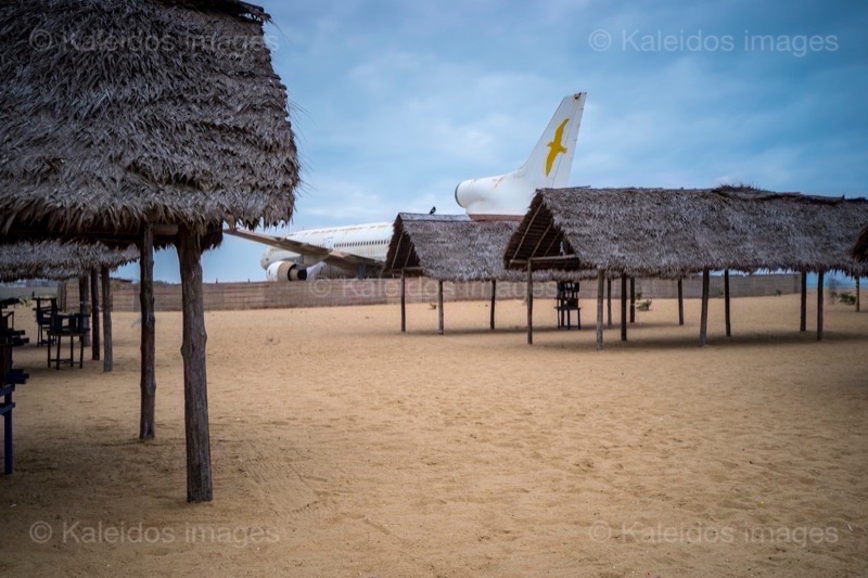 Barakah, L1011 TriStar, Lockheed L1011, abandoned TriStar, aircraft wreck, grounded aircraft, white fuselage, derelict cockpit, empty cabin, Cotonou beach, Cadjéhoun beach, Cotonou Benin, West Africa, Benin airport, Atlantic coastline, coastal Benin, corroded wreck, marine rust, deterioration, salt corrosion, decay, abandonment, dereliction, dilapidation, environmental erosion, aviation heritage, industrial archaeology, failed tourism project, open-air museum, urban art, colonial relic, Benin history, alternative tourism, architectural curiosity, aviation history, hajj pilgrimage, Air Rum airline, defunct carrier, humanitarian transport, international migration, historical traces, industrial heritage, stranded aircraft, tourist attraction, urban exploration, ruin photography, beach adventure, Beninese leisure, amusement park, immersive experience, urban safari, overcast sky, sunset beach, tropical storm, coastal erosion, sea salt, Atlantic Ocean, white sand, coastal vegetation, gleaming white, nature-machine contrast, aerospace geometry, outstretched wings, empty windows, cabin stairway, cockpit instruments, time displacement, obsolescence, suspended time, liminality, aborted transformation, urban memory, unfinished dream, nature-industry symbiosis, tourism ghost, frozen history